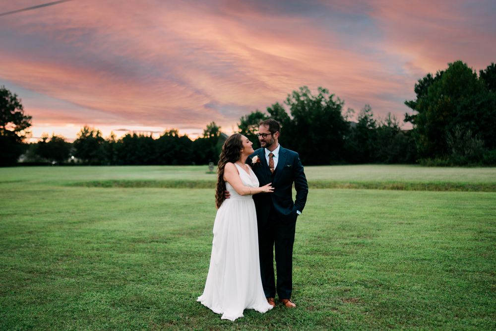 The newlyweds embrace in an open field beneath a pink sunset.