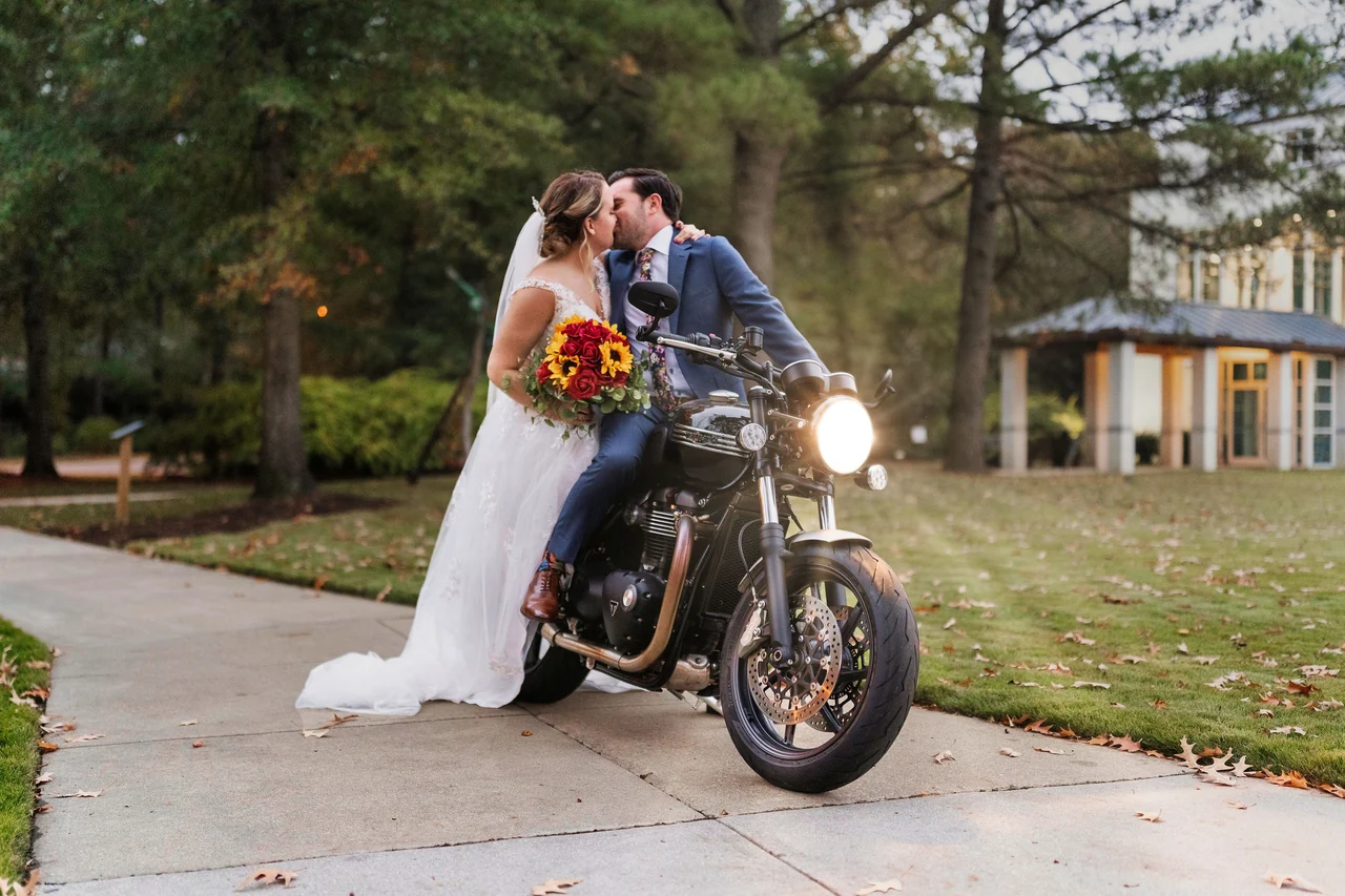 Bride and groom kiss beside a motorcycle outside a wooded venue.