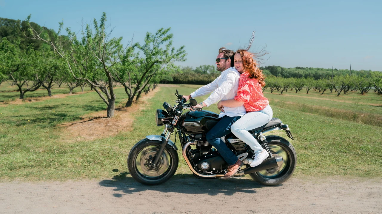 Couple rides a motorcycle through a green orchard.