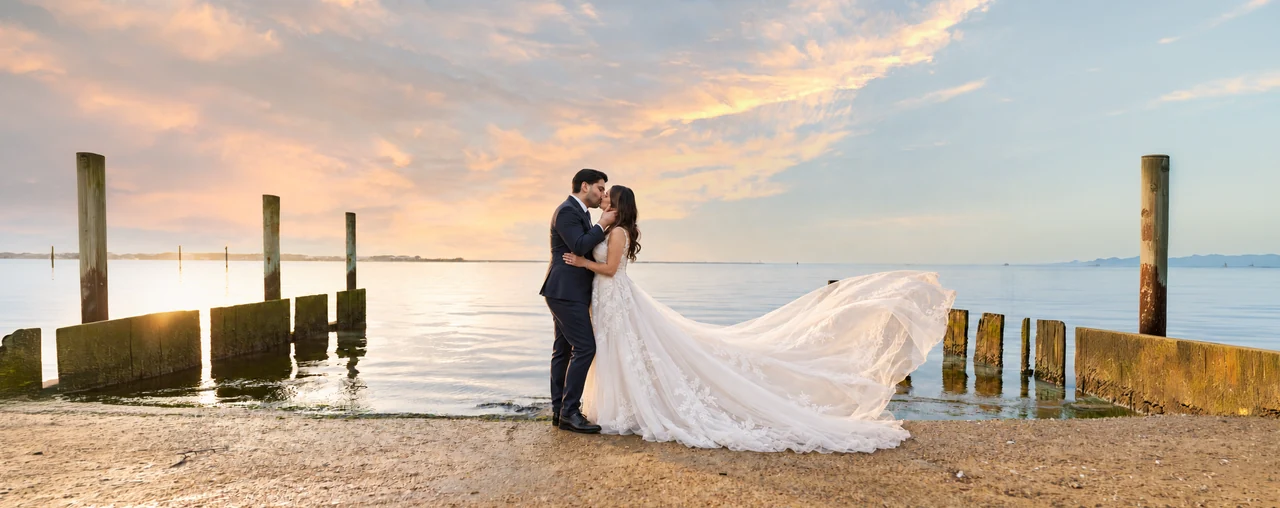 Newlyweds kiss on a small waterfront beach while the wedding dress flows to the right under a pastel sunset.