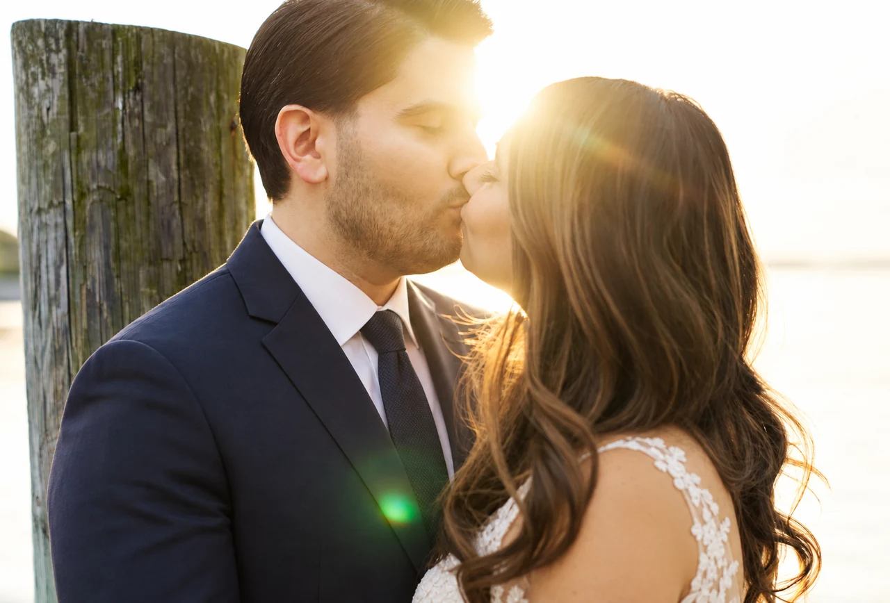 Newlyweds kiss beside waterfront pilings in golden backlight.