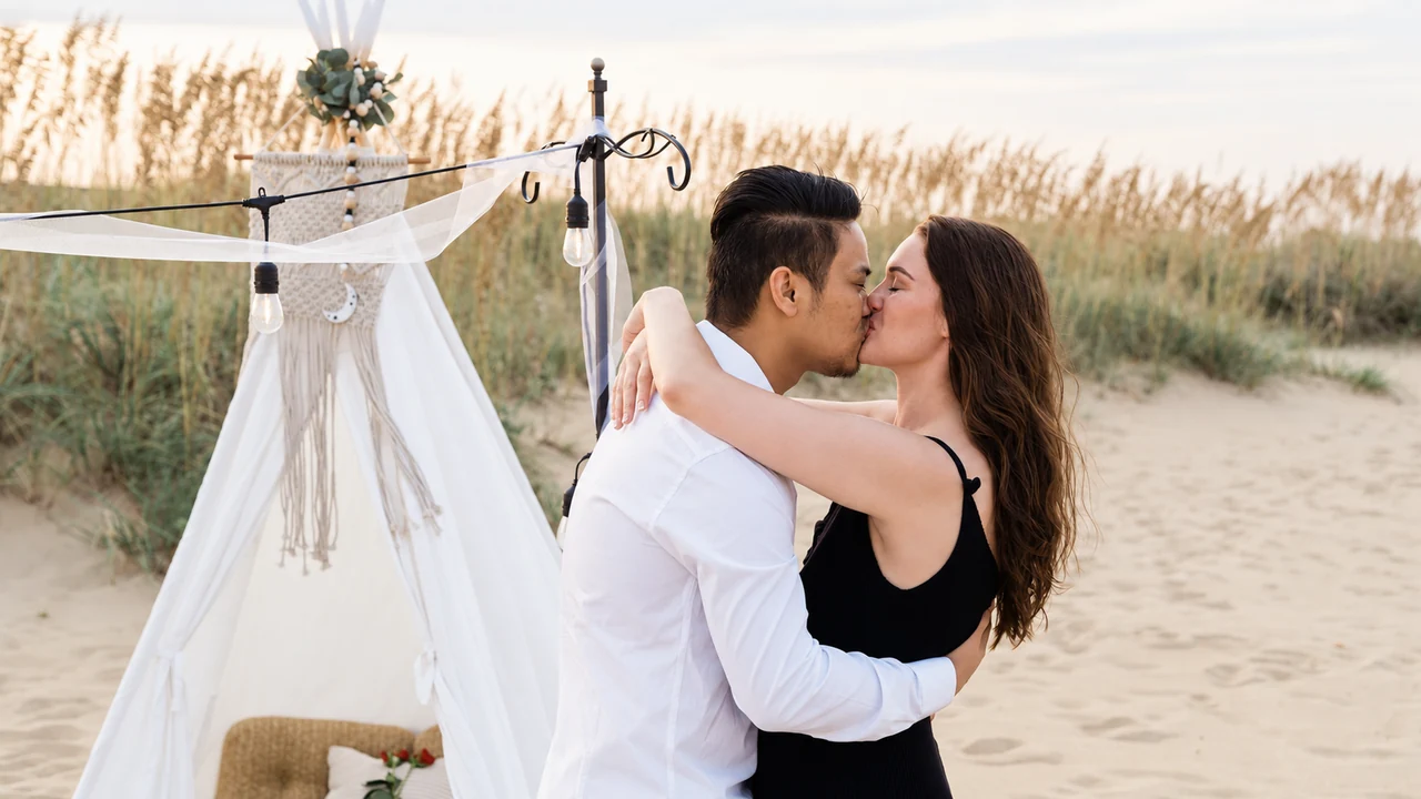 Couple kisses beside a styled beach picnic and sea grass.