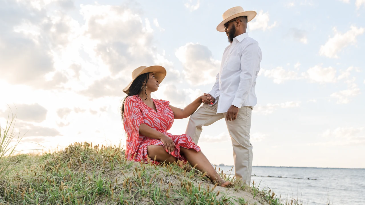 Couple in coordinated light clothing and hats hold hands on a grassy dune against the sky.