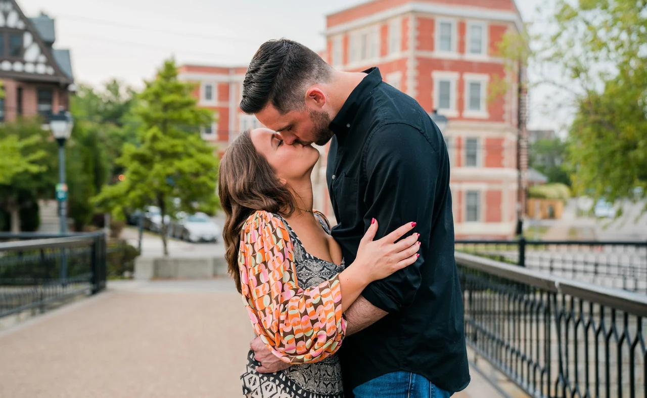 Couple kisses beside an iron railing with brick buildings behind them.