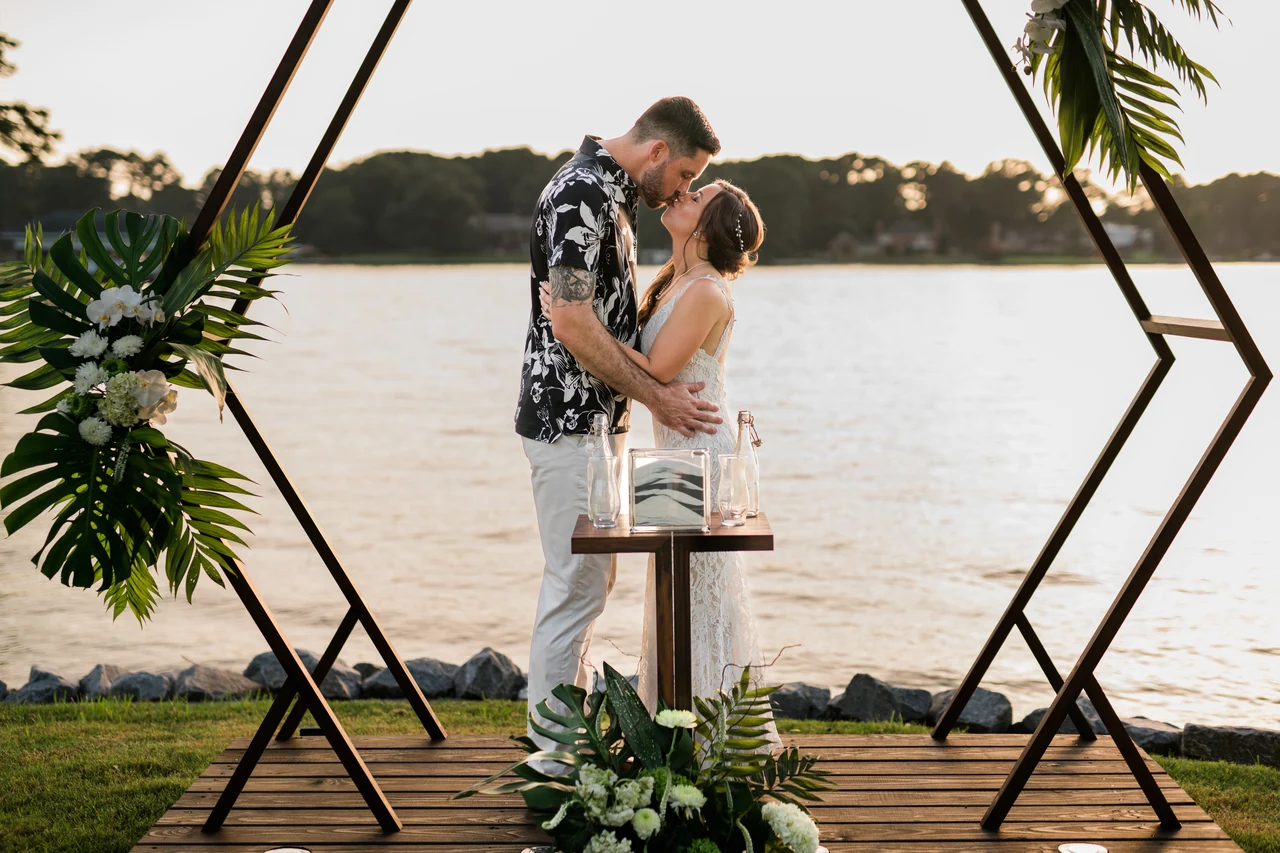 Newlyweds kiss beneath a geometric wooden arch beside the water.