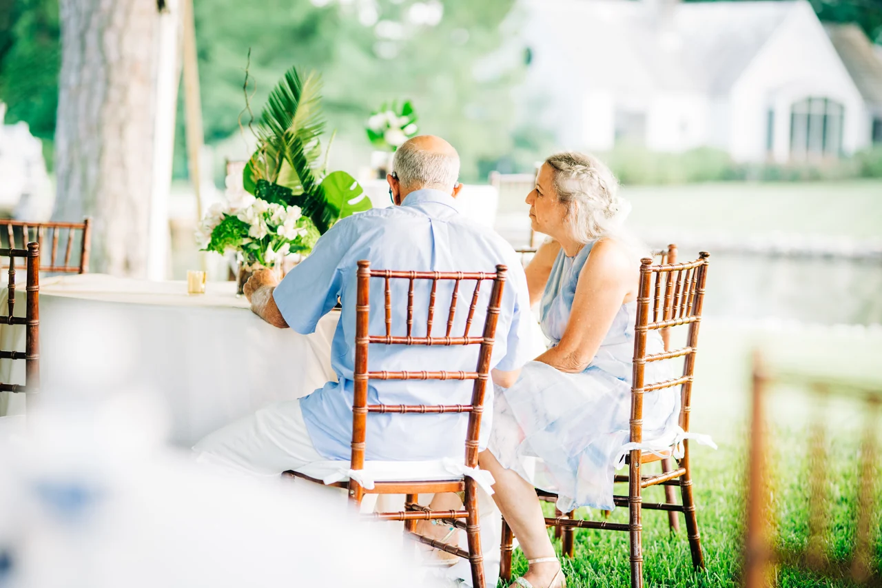 Two older guests sit at a reception table looking out toward the water.