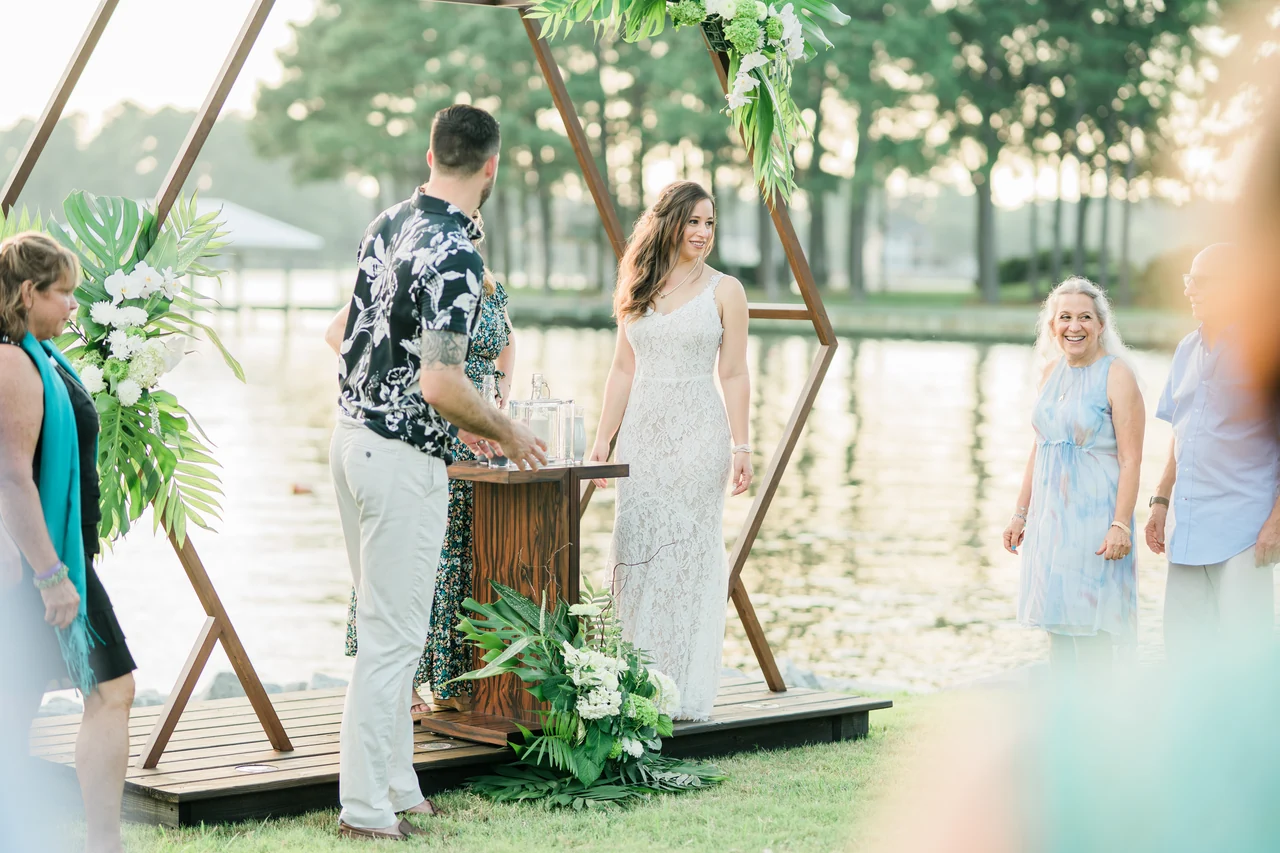 Bride and groom stand at a waterside geometric arch with guests nearby.