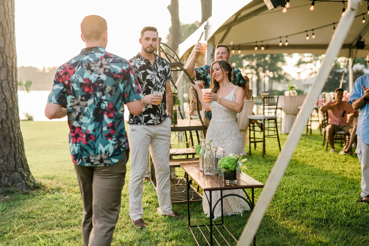 Bride and groom chat with guests on the waterfront lawn beside reception tables.