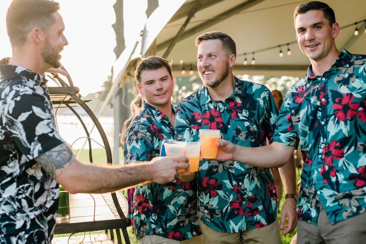 Groom and groomsmen in tropical shirts toast with colorful drinks.