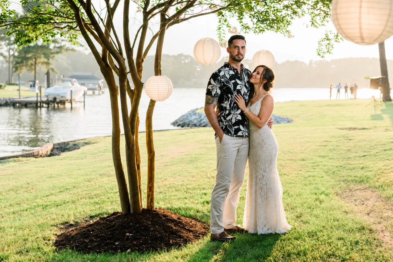Newlyweds stand under a tree and hanging lanterns on the waterfront lawn.