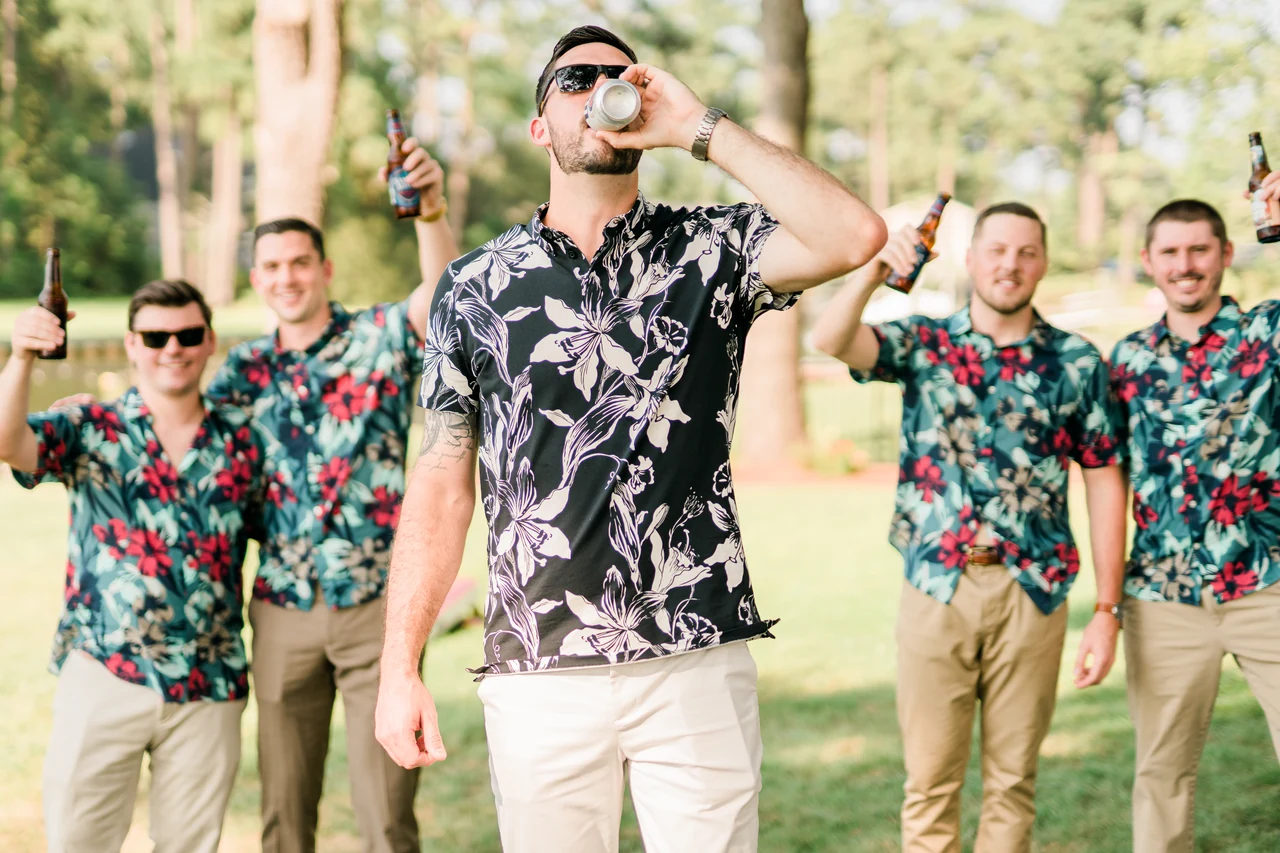 Groom and groomsmen raise drinks and celebrate on a sunny lawn.
