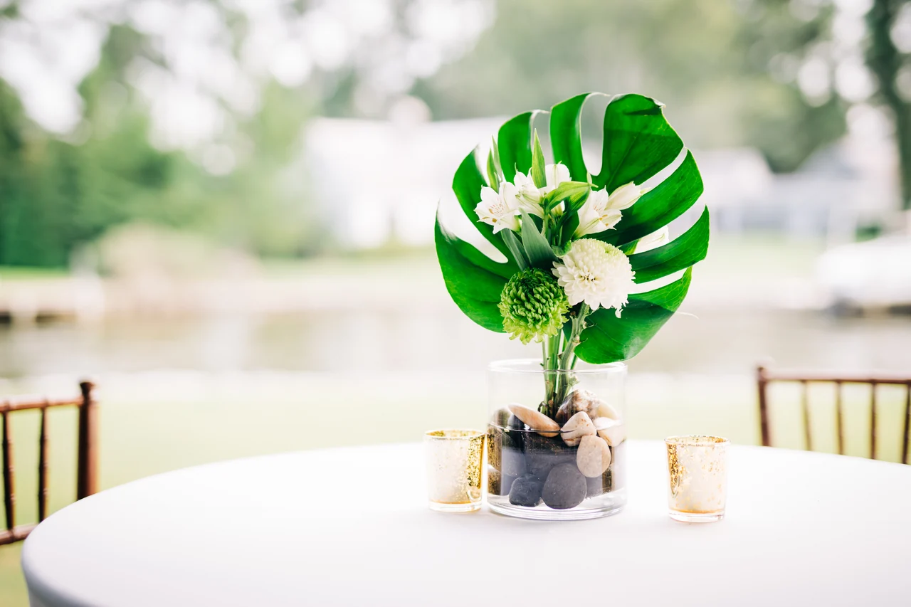 Tropical leaf and white floral centerpiece on a round reception table beside the water.