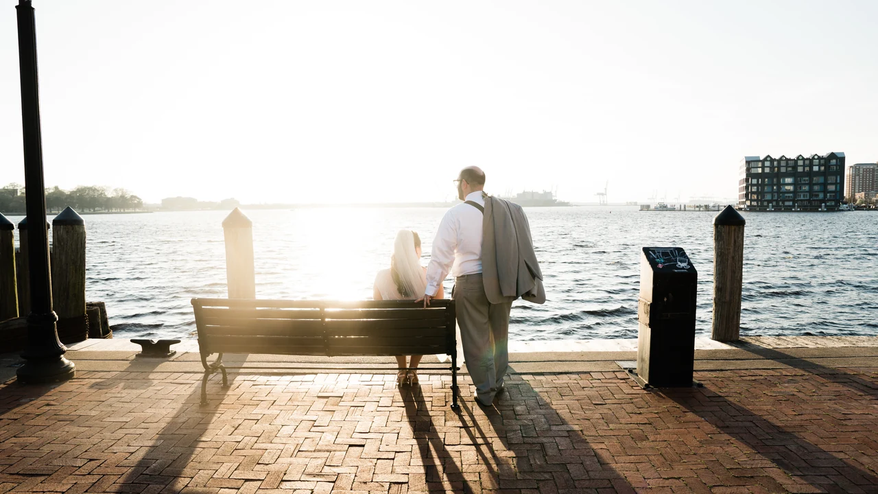 Couple pauses at a waterfront bench in bright low sun on a brick promenade.