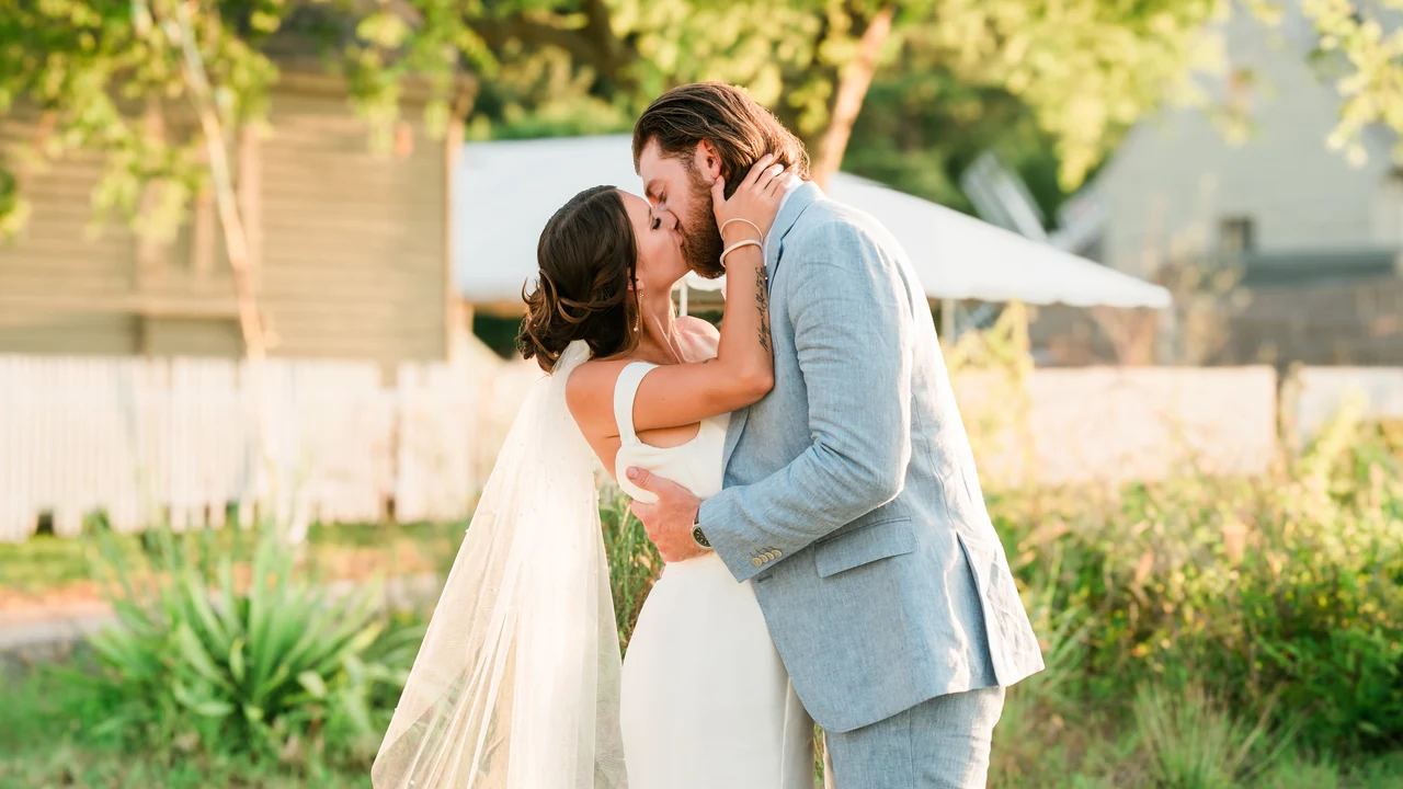 Newlyweds kiss in golden light beside a garden and pale fence.