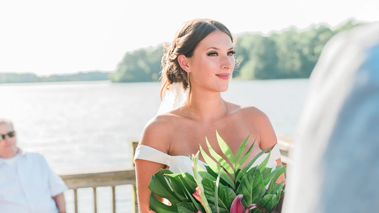 Bride holds a tropical bouquet and looks toward the groom by the water.