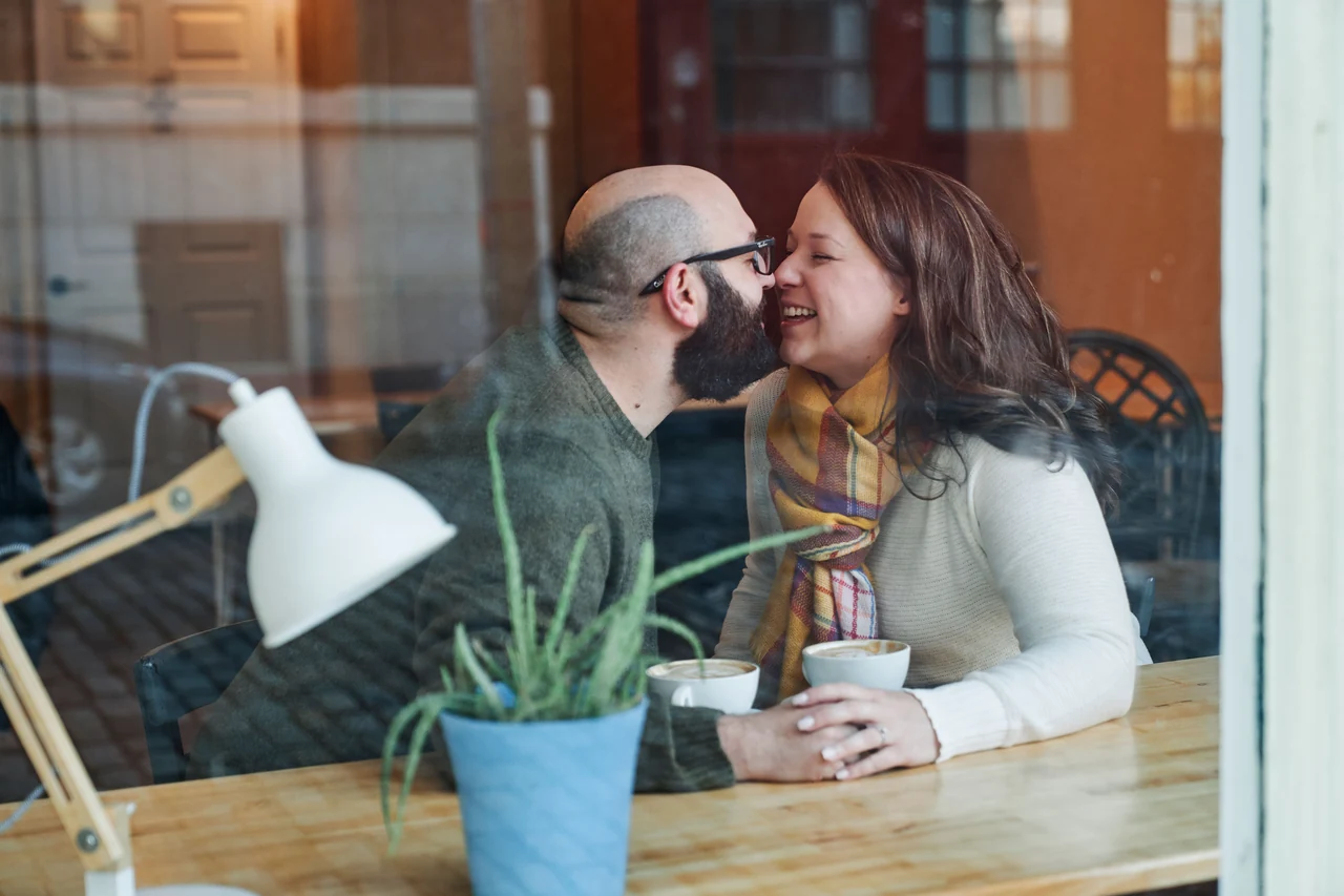 Couple shares an affectionate moment at a cafe table, photographed through a window.