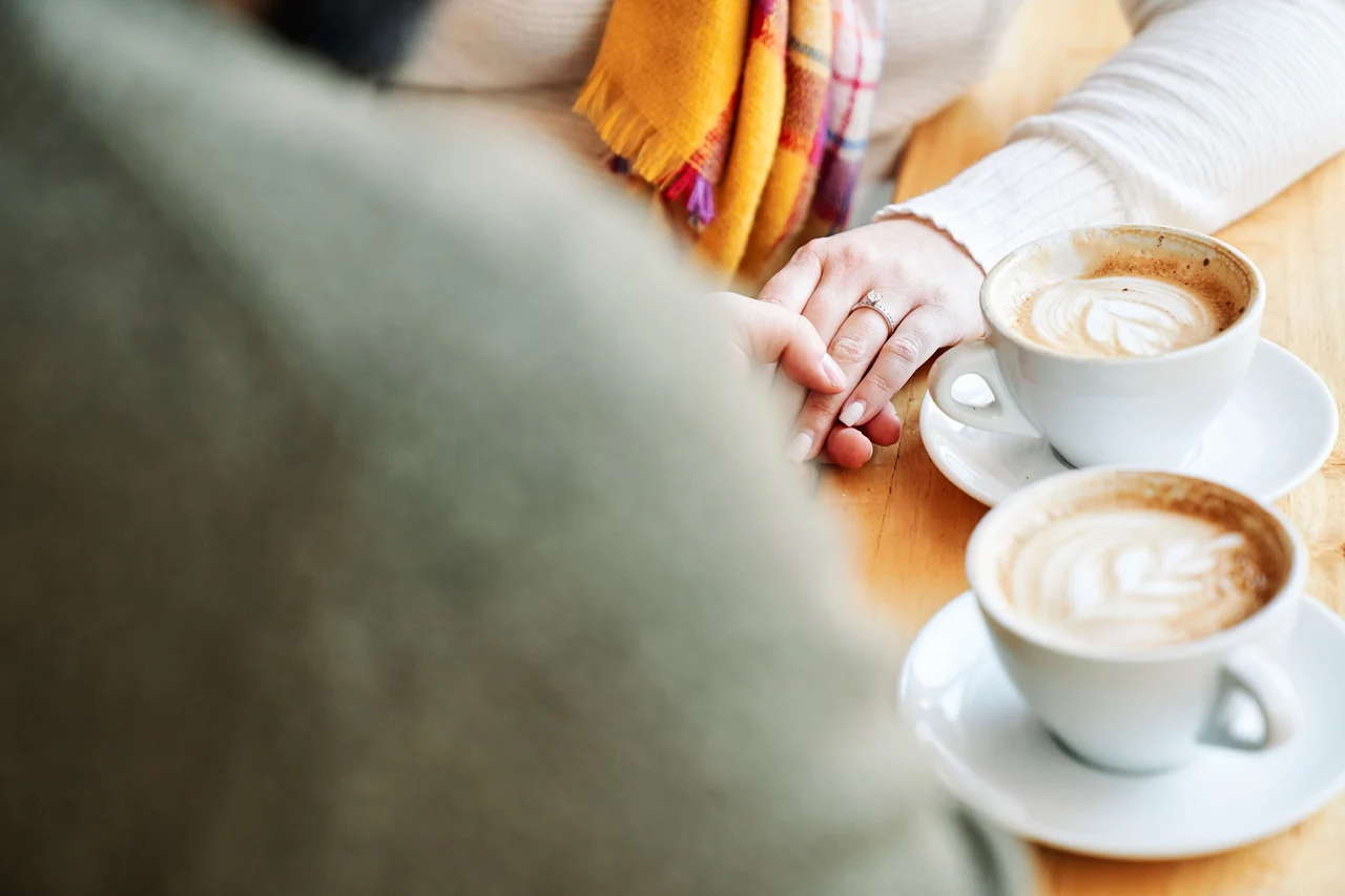Close view of linked hands with an engagement ring beside two cappuccinos.