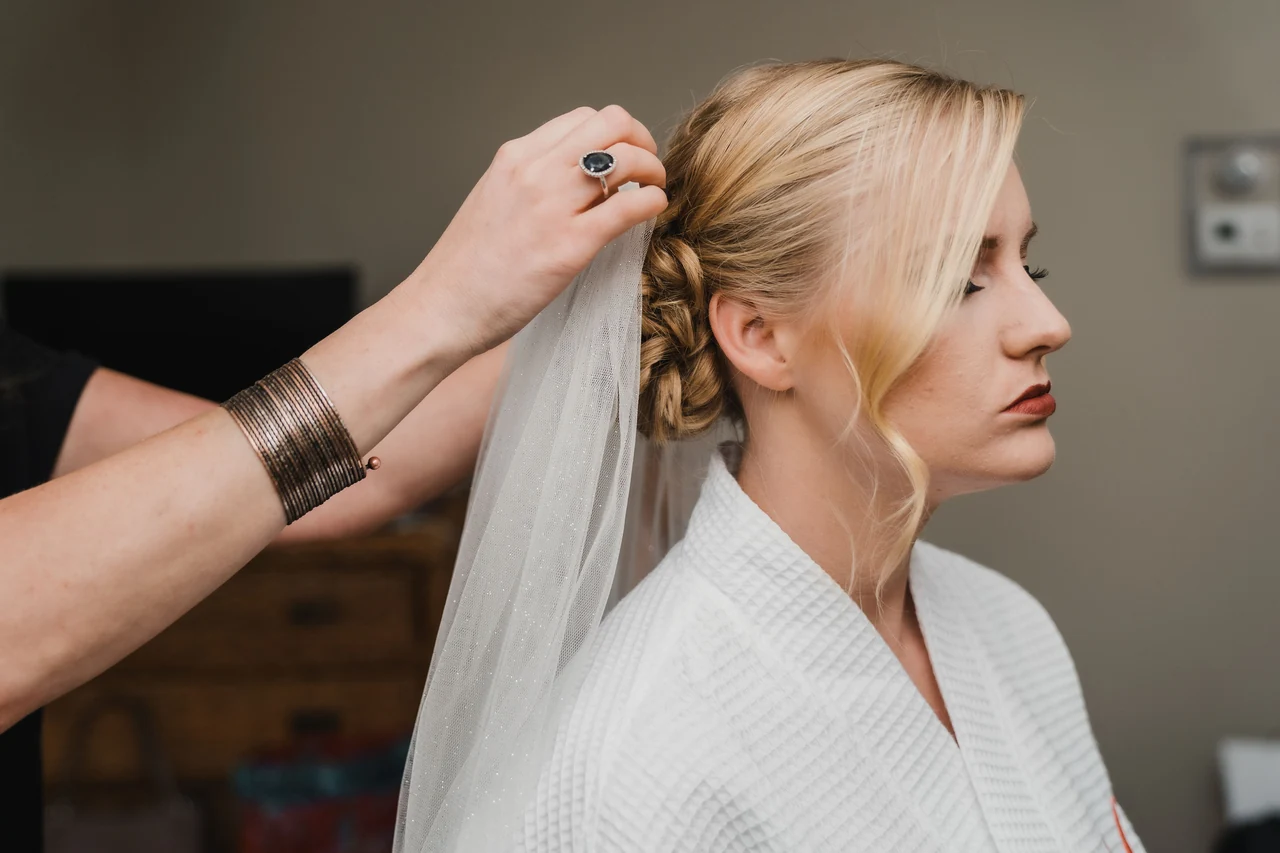 Bride has her braided hair and veil adjusted while wearing a white robe.