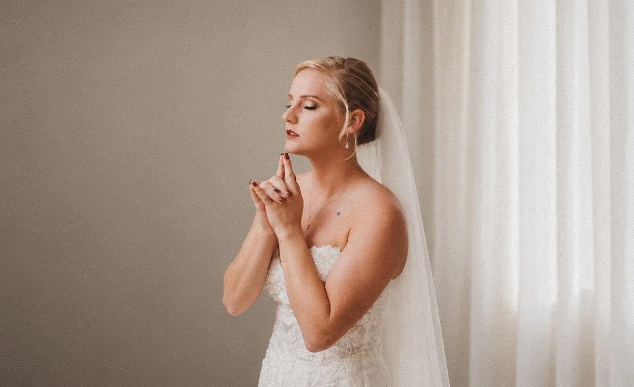 Bride in a lace wedding dress pauses beside a softly lit window with her hands beneath her chin.