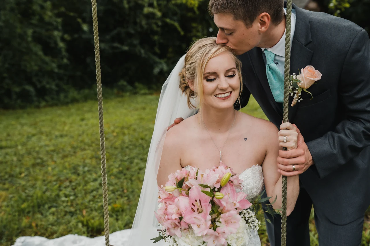 Groom kisses seated bride on the forehead while she holds a pink bouquet on a garden swing.
