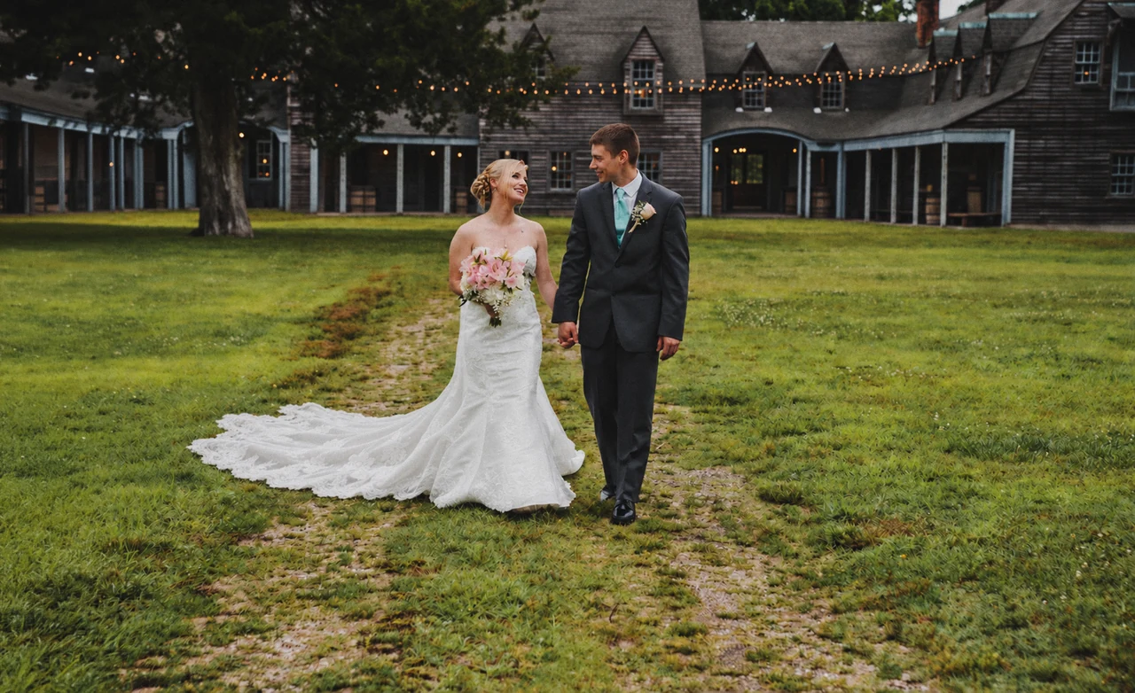 Newlyweds walk hand in hand across a green lawn in front of a weathered shingle venue and string lights.