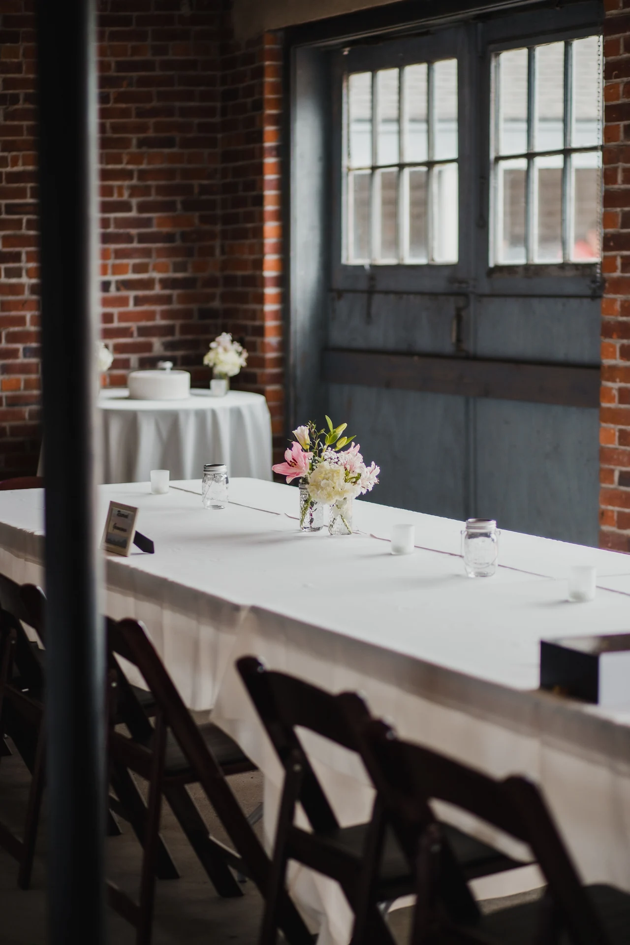 Quiet reception tables with small flower arrangements beside brick walls and tall windows.